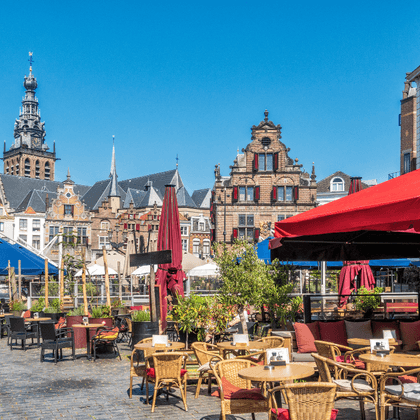 Outdoor café terraces with colorful umbrellas on Grote Markt square in Nijmegen. Historic Dutch buildings and a church tower under blue sky.