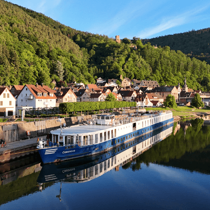 Blau-weißes Flusskreuzfahrtschiff Casanova am Kai eines Dorfes mit traditionellen Häusern, grünen bewaldeten Hügeln und ruhigen Wasserspiegelungen.
