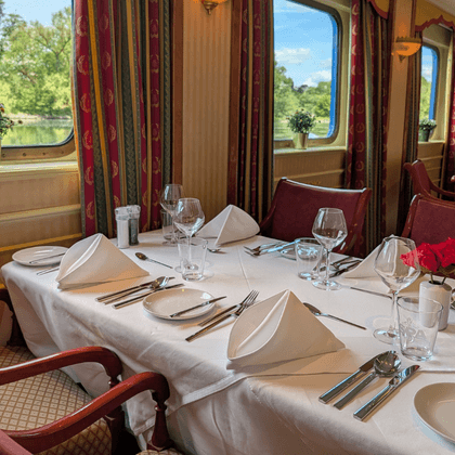 Elegant restaurant aboard Casanova with white-clothed tables, burgundy chairs, and large windows overlooking a green riverbank.