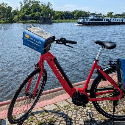 Rotes Fahrrad mit blauen Taschen am Flussufer geparkt. Ein Passagierschiff ist auf dem Wasser sichtbar mit Bäumen am gegenüberliegenden Ufer.