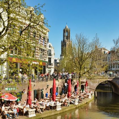 Canal-side cafes in Utrecht with Dom Tower Outdoor dining terraces along Utrecht canal with red umbrellas. Historic buildings line the waterway with Dom Tower visible in background.