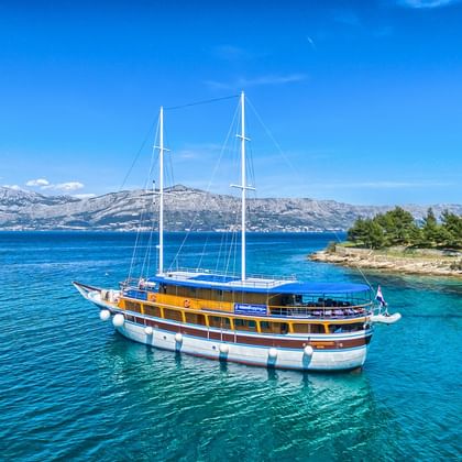 San Snova, eine traditionelle hölzerne Segelyacht mit zwei Masten, vor Anker in kristallklarem türkisfarbenem Wasser nahe einer felsigen Küste.