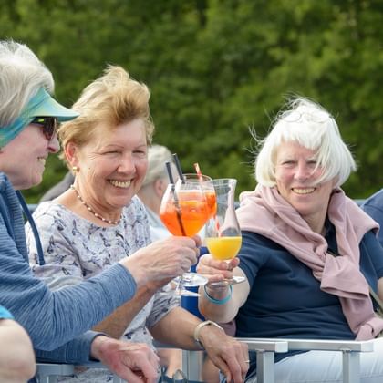 Four elderly passengers sitting together on a cruise ship deck, toasting with colorful cocktails. Green foliage is visible in the background.
