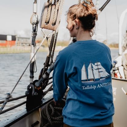 Crew-Mitglied in blauem Sweatshirt mit Tallship Antigua Logo steht an der Takelage und blickt über das Wasser im Hafen.