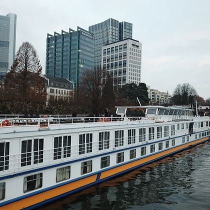 White river cruise ship MS Classica with blue and orange trim moored on a river, with modern glass buildings and bare trees in the background.
