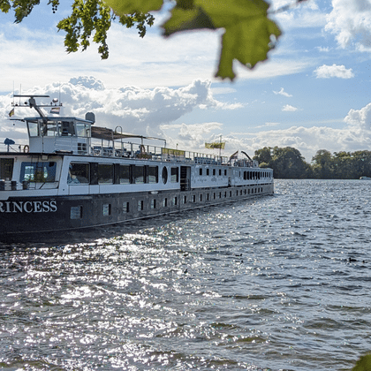Weißes Flusskreuzfahrtschiff namens Princess an einem Holzsteg an einem sonnigen Tag. Das Schiff ist von grünen Blättern eingerahmt mit See und bewaldeter Küste.