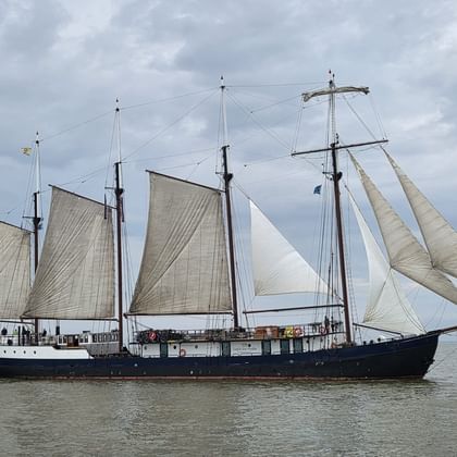 Four-masted sailing ship Leafde fan Fryslân with dark hull and cream-colored sails on calm water under cloudy sky.