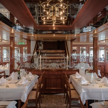 Elegant ship restaurant with white-clothed tables, wooden chairs, and a historic paddle wheel centerpiece. Mirrored ceiling and warm lighting.