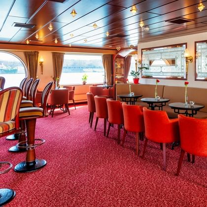 Interior of MS Switzerland salon bar with red carpet, orange chairs, round tables, bar stools, and large windows overlooking the water.