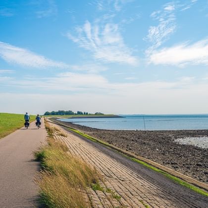 Two cyclists on paved path along coastal dike in Zeeland. Green grass slopes meet rocky shoreline under blue sky with wispy clouds.