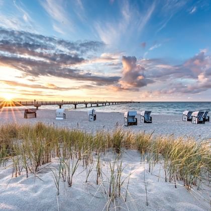 Sandy beach on Rügen with beach chairs and dune grass in foreground. A wooden pier extends into the Baltic Sea under a dramatic sunset sky.