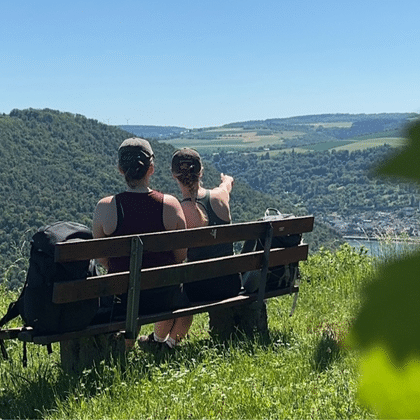 Zwei Personen sitzen auf Holzbank mit Blick auf grüne Hügel und Flusstal mit Stadt. Sonniger Tag mit blauem Himmel und üppiger Vegetation.