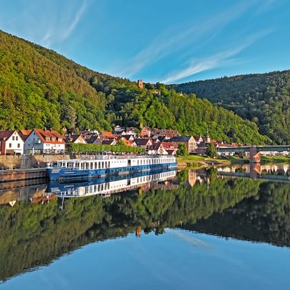 River cruise ship Casanova moored along a riverside village with colorful houses, forested hills, and perfect water reflections under blue sky.