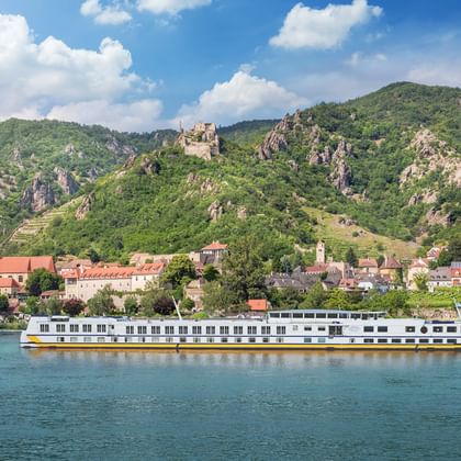 Weißes Flusskreuzfahrtschiff MS Vista Classica auf der Donau vor historischer Stadt mit Kirchtürmen und grünen Bergen mit Burgruine.
