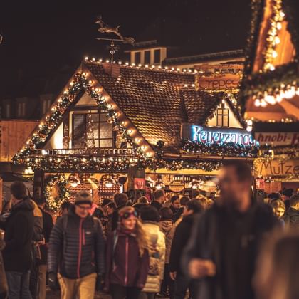 Belebter Weihnachtsmarkt bei Nacht mit beleuchteten Holzbuden und geschmücktem Häuschen. Warme Lichter schaffen festliche Atmosphäre.