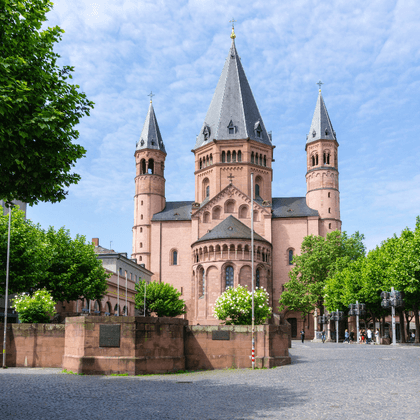 Mainz Cathedral with pink sandstone facade and six towers framed by green trees. The Romanesque church stands on a cobblestone square.