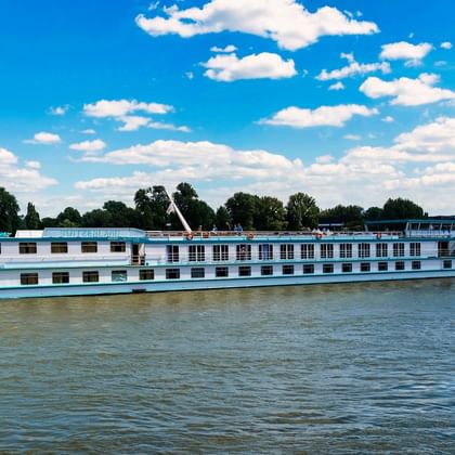 Weiß-blaues Flusskreuzfahrtschiff MS Switzerland auf ruhigem Wasser unter blauem Himmel mit weißen Wolken. Bäume und Gebäude am Ufer sichtbar.
