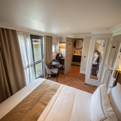 Elegant river cruise suite with double bed, balcony door, seating area, wardrobe, and warm wood flooring on the Magnifique X upper deck.