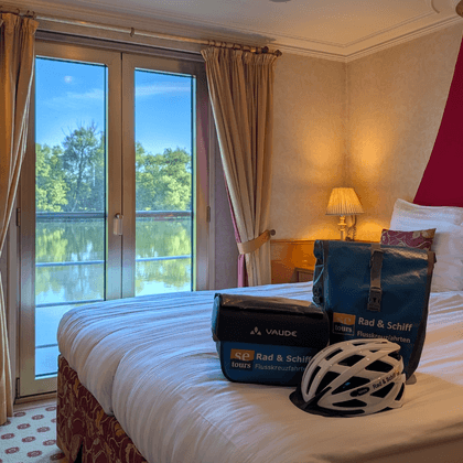 Double cabin on Casanova upper deck with two beds, balcony door, river view, beige walls, burgundy accent wall, and cycling equipment on bed.