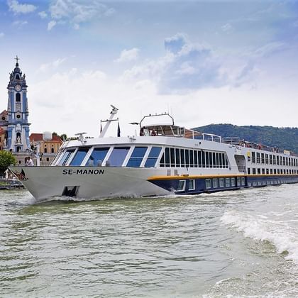 Weißes Flusskreuzfahrtschiff MS SE-MANON fährt auf der Donau vor historischer Stadt mit barockem Kirchturm und roten Dächern.