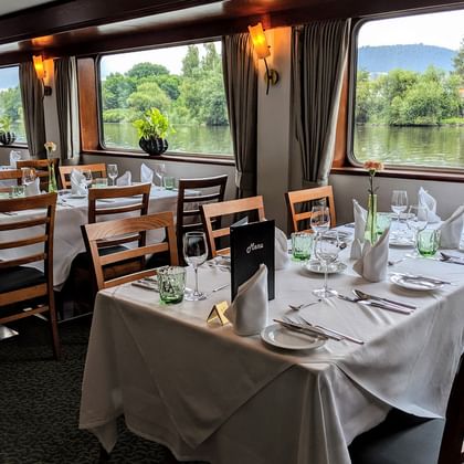 Eleganter Speisesaal an Bord der Classica mit weiß gedeckten Tischen, Holzstühlen und großen Fenstern mit Blick auf Fluss und grüne Landschaft.