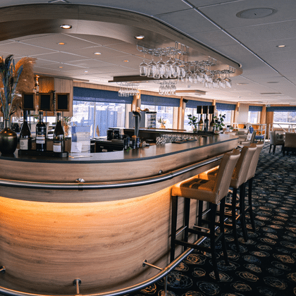 Curved bar with light wood paneling and bar stools on DE Nassau. Wine bottles and glasses visible, with dining area and windows in background.