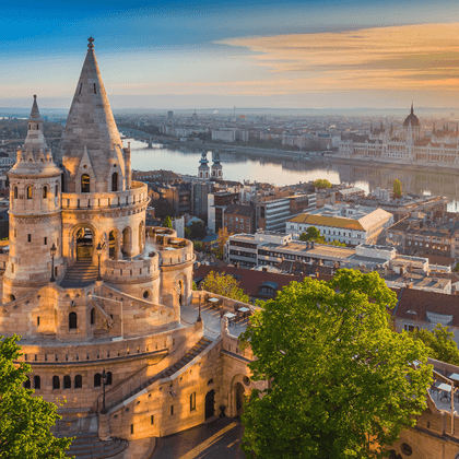 Fisherman's Bastion towers in Budapest at sunrise with the Danube River and Parliament building visible in the background under golden sky.