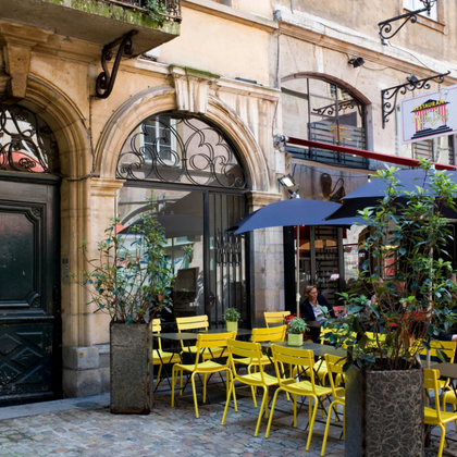 Gelbe Cafémöbel auf einer Kopfsteinpflaster-Terrasse in der Provence, umgeben von historischen Gebäuden mit Bogentüren und Schmiedeeisen.