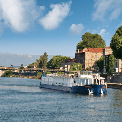 Weiß-blaues Flusskreuzfahrtschiff auf der Rhône bei Tournon mit historischen Gebäuden und Brücke im Hintergrund unter blauem Himmel.