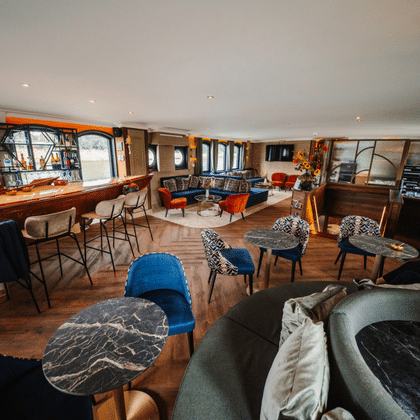 Spacious lounge on Magnifique X with wooden bar, blue velvet chairs, patterned seating, red armchairs, and elegant chandelier. Large windows line the walls.