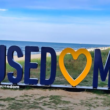 Large blue and orange USEDOM letters with heart symbol on sandy beach. Baltic Sea and dune grass visible in background under blue sky.