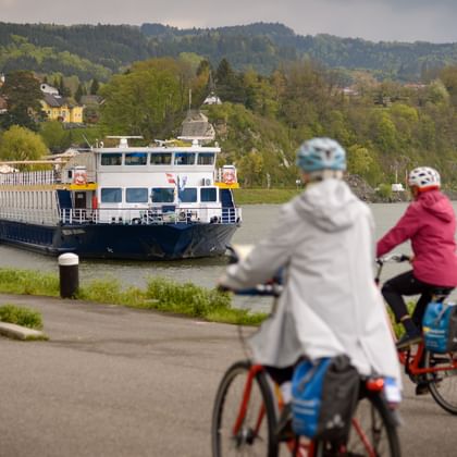 Zwei Radfahrer mit Helmen beobachten die MS Prinzessin Katharina Fähre am Pier. Hügelige Landschaft und Häuser im Hintergrund.