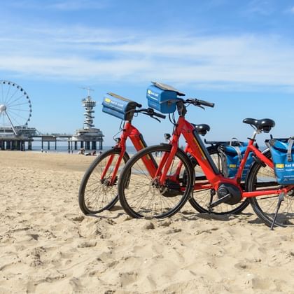 Zwei Leihfahrräder am Sandstrand von Scheveningen mit Pier und Riesenrad im Hintergrund unter blauem Himmel mit Wolken.