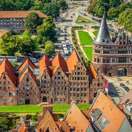 Luftaufnahme des Lübecker Holstentors mit Doppeltürmen und historischen Backsteingebäuden mit orangen Dächern. Grüne Rasenflächen und baumgesäumte Straßen sichtbar.