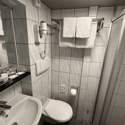 Compact bathroom on Classica ship with white tiles, sink, toilet, glass shower, mirror, towels, and toiletries on black counter.