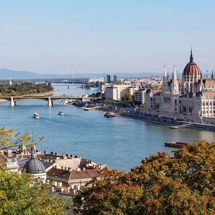 Panoramablick auf das Parlamentsgebäude in Budapest mit seiner markanten roten Kuppel am Donauufer, eingerahmt von Herbstbäumen und Brücken.