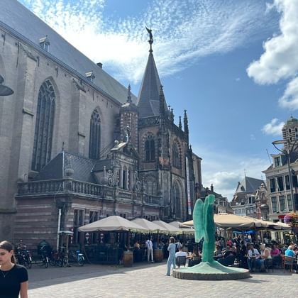 Gothic church with tall spire dominates a cobblestone square in Zwolle. Outdoor café terraces with umbrellas line historic buildings under blue sky.