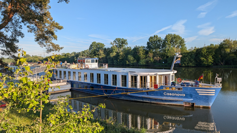 Blau-weißes Flusskreuzfahrtschiff Casanova an einem Holzsteg auf ruhigem Fluss, umgeben von grünen Bäumen unter blauem Himmel.