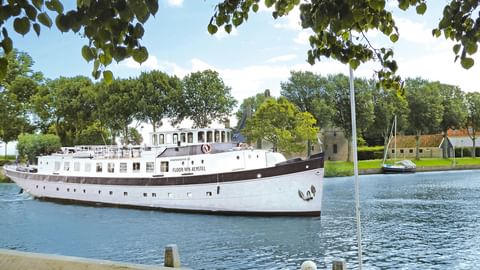 Weißes Passagierschiff Floor van Aemstel mit dunklem Rumpf auf Wasserstraße, eingerahmt von Baumzweigen. Historische Gebäude im Hintergrund.