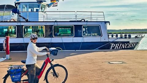 Cyclist with red e-bike and helmet standing on dock next to MS Princess ship. Blue and white vessel moored on calm water under cloudy sky.