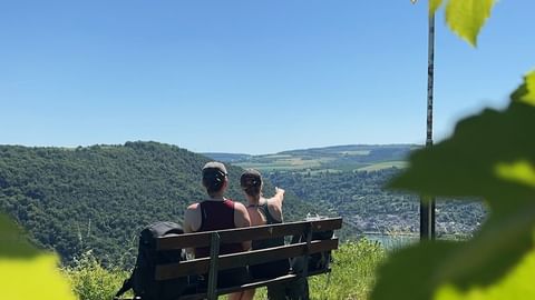 Zwei Wanderer sitzen auf einer Holzbank mit Blick auf Oberwesel und das Rheintal, umgeben von grünen Hügeln und Weinbergen unter blauem Himmel.