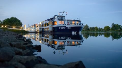 MS Princess cruise ship moored at dock during evening twilight, with warm lights glowing from windows and perfect reflection in calm water.