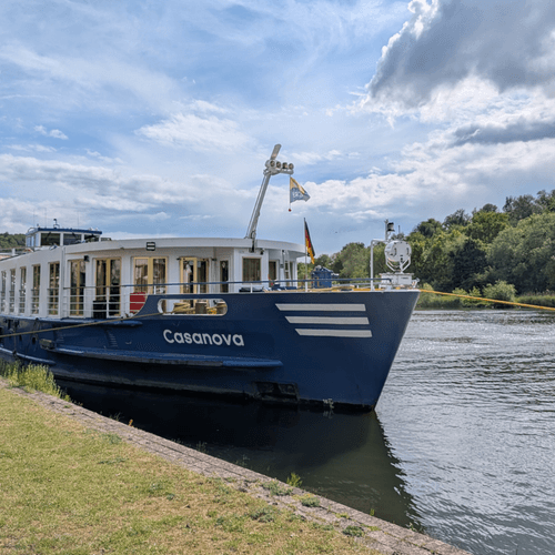 River cruise ship Casanova with dark blue hull and white upper deck moored at grassy riverbank under cloudy sky with green trees in background.