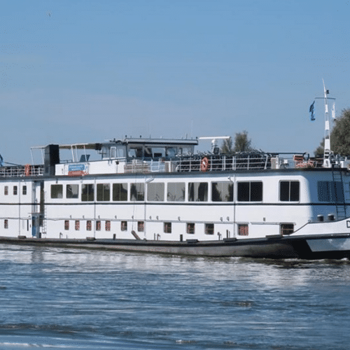 White river cruise ship De Nassau sailing on calm water. The multi-deck vessel has large windows and a sun deck, with green riverbanks visible.