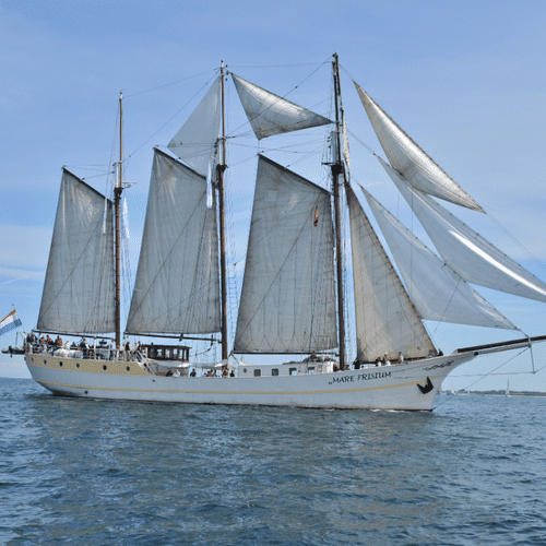 White three-masted sailing ship Mare Frisium with gray sails on calm blue water under clear sky, flying the Dutch flag.
