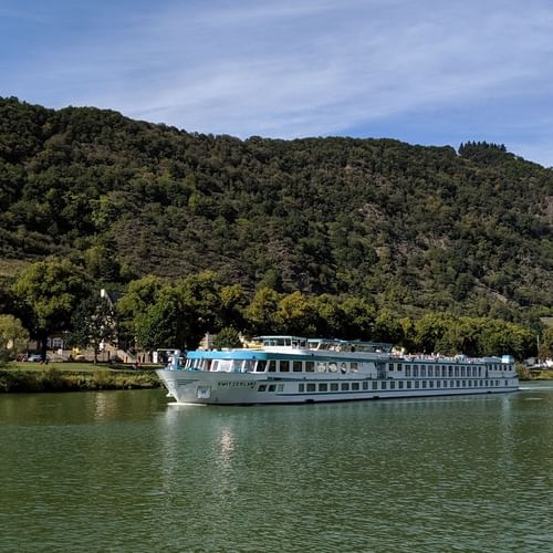 Weißes Flusskreuzfahrtschiff Switzerland auf ruhigem grünen Wasser mit bewaldeten Hügeln und kleinem Dorf im Hintergrund unter blauem Himmel.
