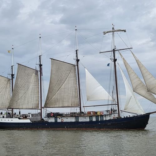 Four-masted sailing ship Leafde fan Fryslân with dark hull and cream-colored sails on calm water under cloudy sky.