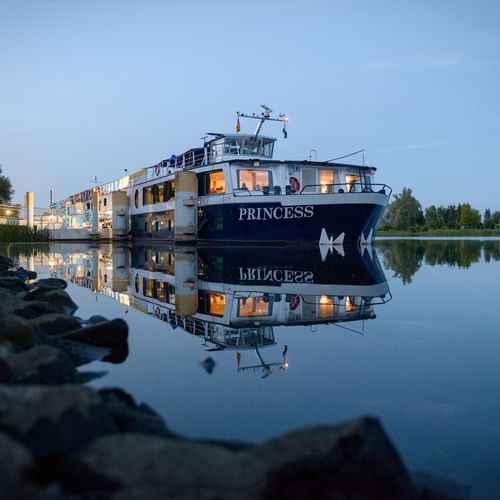 MS Princess cruise ship moored at dock during evening twilight, with warm lights glowing from windows and perfect reflection in calm water.