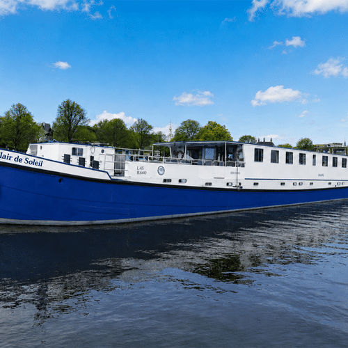 Flusskreuzfahrtschiff Clair de Soleil mit blauem Rumpf und weißem Aufbau am Kai. Bäume und Gebäude im Hintergrund unter blauem Himmel.