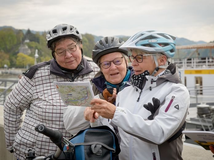 Drei ältere Radfahrer mit Helmen betrachten gemeinsam eine Karte in der Nähe eines Hafens mit Booten im Hintergrund.